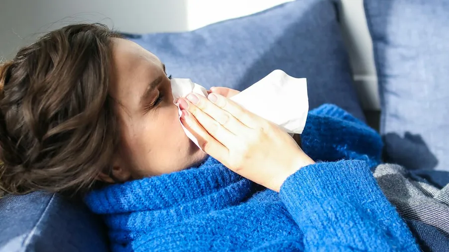 Žena v modrém svetru leží na gauči a smrkáním do kapesníku se snaží ulevit od rýmy